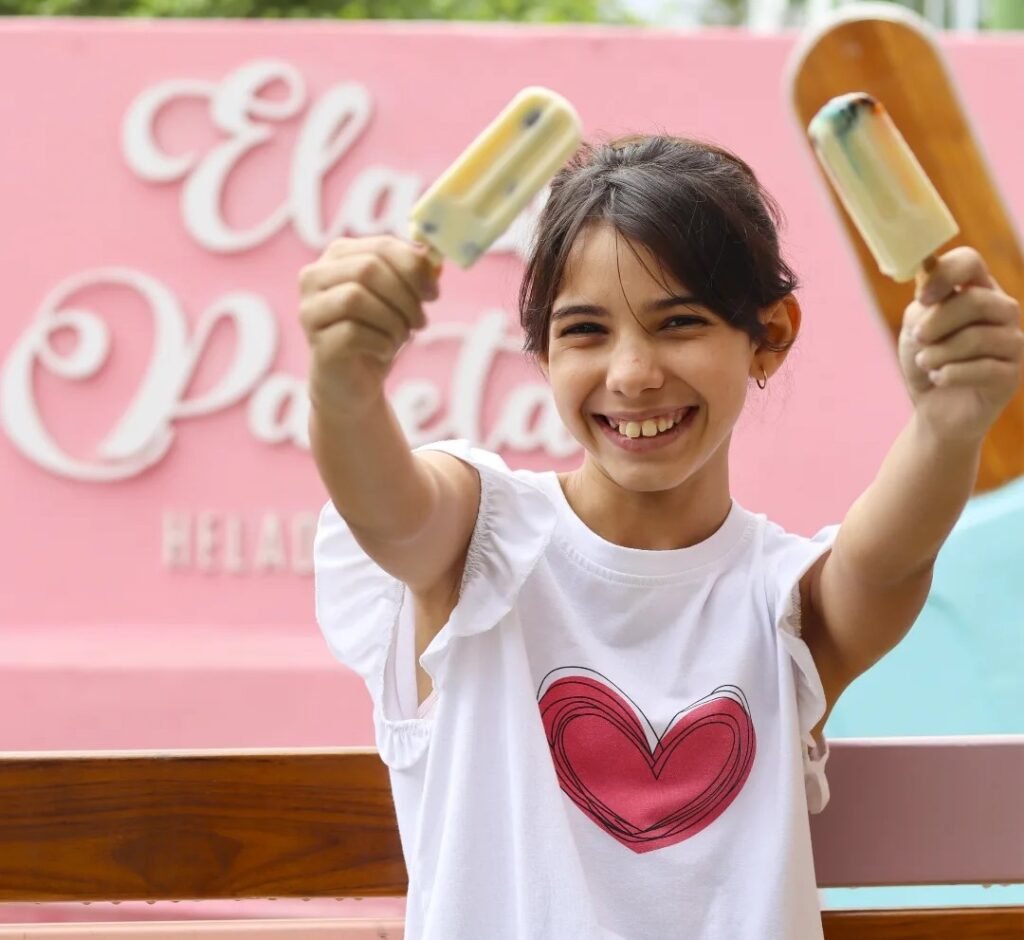 niña sonriendo con dos paletas de la helado en sus manos con logo de ela & paleta en el fondo